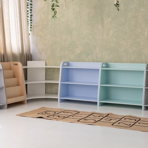 May include: Four wooden bookshelves in a playroom setting. Two shelves are light blue, one is light green, and one is white. The shelves have three shelves each. A brown and beige rug with black numbers is on the floor in front of the shelves.