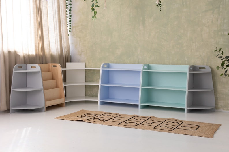 May include: Four wooden bookshelves in a playroom setting. Two shelves are light blue, one is light green, and one is white. The shelves have three shelves each. A brown and beige rug with black numbers is on the floor in front of the shelves.