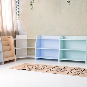 May include: Four wooden bookshelves with different colored fronts. The shelves are arranged in a row against a wall. The shelves are empty. A brown and black hopscotch mat is in front of the shelves.