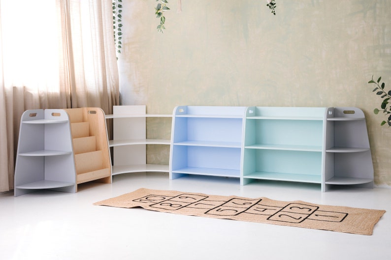 May include: Four wooden bookshelves with different colored fronts. The shelves are arranged in a row against a wall. The shelves are empty. A brown and black hopscotch mat is in front of the shelves.