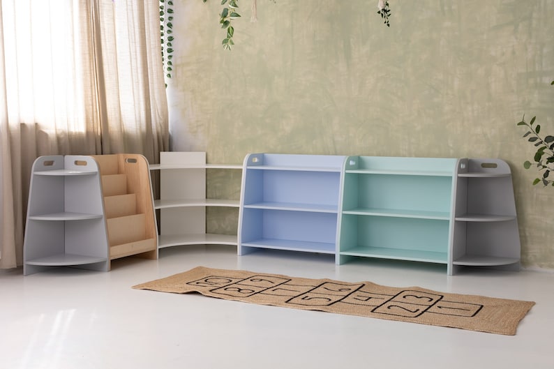 Puede incluir: Cuatro estanter&iacute;as de madera en un ambiente de sala de juegos. Dos estanter&iacute;as son de color azul claro, una es de color verde claro y una es blanca. Las estanter&iacute;as tienen tres estantes cada una. Una alfombra marr&oacute;n y beige con n&uacute;meros negros est&aacute; en el suelo delante de las estanter&iacute;as.
