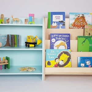 May include: Two wooden bookshelves, one light blue and one natural wood, with different sized shelves. The blue bookshelf has a stuffed duck, a wooden toy truck, and a wooden toy train on the shelves. The natural wood bookshelf has several children's books on the shelves, including "Za Duzo Marchewek" and "Pugio".