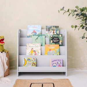 May include: A gray wooden bookshelf with five shelves holds a collection of children's books with colorful covers. The books are in Polish and feature illustrations of animals, people, and objects. A green dinosaur plush toy sits in a wicker basket on the left side of the image.