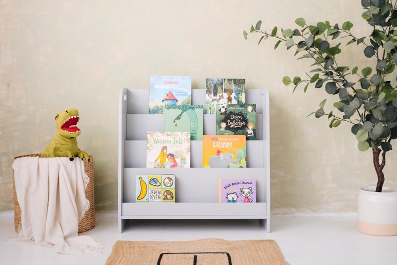 May include: A gray wooden bookshelf with five shelves holds a collection of children's books with colorful covers. The books are in Polish and feature illustrations of animals, people, and objects. A green dinosaur plush toy sits in a wicker basket on the left side of the image.