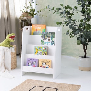 Pu&ograve; includere: Libreria in legno bianco con quattro ripiani, che contiene una variet&agrave; di libri per bambini. La libreria &egrave; in piedi su un pavimento bianco con un tappeto marrone di fronte ad essa. C'&egrave; un dinosauro di peluche verde in un cesto di vimini a sinistra della libreria.