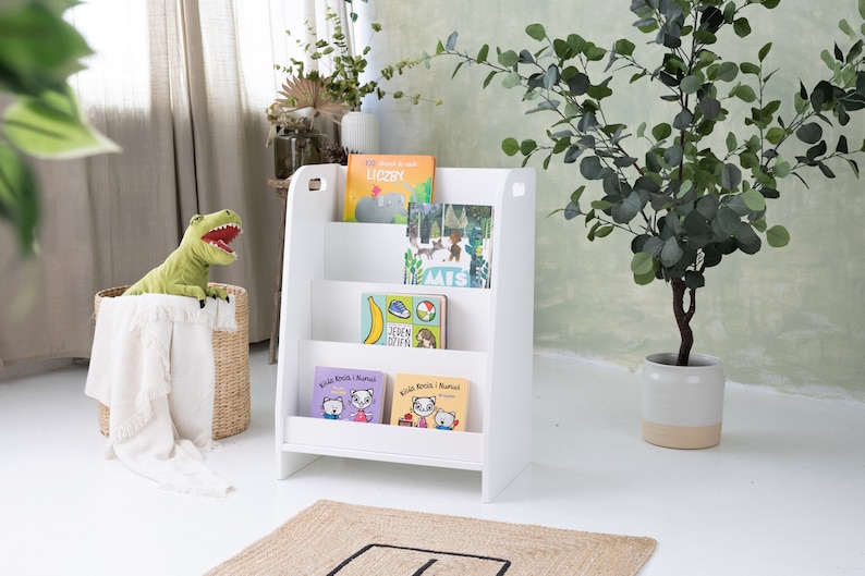 Pu&ograve; includere: Libreria in legno bianco con quattro ripiani, che contiene una variet&agrave; di libri per bambini. La libreria &egrave; in piedi su un pavimento bianco con un tappeto marrone di fronte ad essa. C'&egrave; un dinosauro di peluche verde in un cesto di vimini a sinistra della libreria.