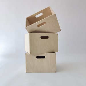 May include: Three stacked, light-colored wooden storage boxes. Each box has a rectangular shape with a handle cut-out on the front. The top box is angled, revealing the interior. The boxes are made of light-colored wood.