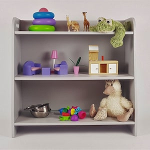May include: A gray wooden shelf unit with three shelves. The top shelf has a green frog plush toy, a stack of colorful rings, and two giraffe figurines. The middle shelf has a purple couch, two chairs, a lamp, a plant, and a wooden toy house. The bottom shelf has a metal bowl, a duck plush toy, and a pile of colorful plastic toys.