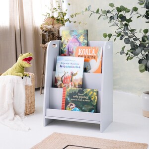 May include: A gray wooden bookshelf with three shelves, holding a variety of children's books. The bookshelf is standing on a light wood floor. A stuffed dinosaur toy is sitting in a wicker basket in the foreground.