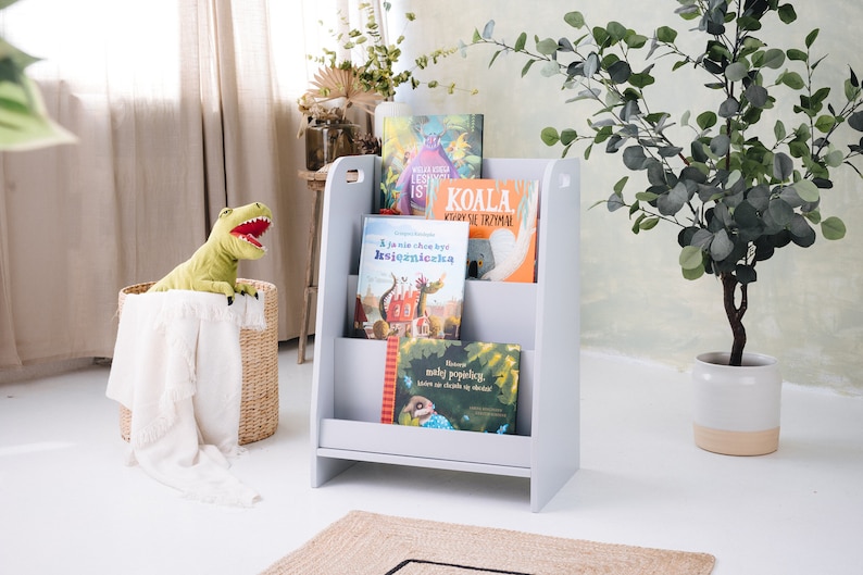 May include: A gray wooden bookshelf with three shelves, holding a variety of children's books. The bookshelf is standing on a light wood floor. A stuffed dinosaur toy is sitting in a wicker basket in the foreground.