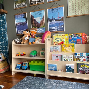 Pu&ograve; includere: Una libreria in legno con due ripiani a sinistra e tre a destra. I ripiani sono pieni di libri per bambini, giocattoli e animali di peluche. I libri includono titoli come "Noisy Diggers", "Construction Site", "Love Trucks", "Kindness Makes Us Strong", "Audubon Trucks" e "Paco le Jazz". I giocattoli includono un camion giocattolo verde, un camion giocattolo rosso e una scatola di giocattoli verde.