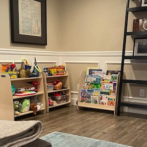 Puede incluir: Dos estanter&iacute;as de madera para juguetes con varios juguetes y libros. Las estanter&iacute;as son de color marr&oacute;n claro y tienen tres estantes cada una. El estante superior de la estanter&iacute;a de la izquierda tiene un coche de juguete rojo, un cami&oacute;n de juguete azul, una pala de juguete amarilla y un casco de juguete plateado. El estante del medio tiene una pelota de juguete azul, un piano de juguete verde y un taladro de juguete plateado. El estante inferior tiene un animal de peluche verde, un animal de peluche marr&oacute;n y un coche de juguete azul. El estante superior de la estanter&iacute;a de la derecha tiene una variedad de libros coloridos. El estante del medio tiene una variedad de libros coloridos. El estante inferior tiene una variedad de libros coloridos. Las estanter&iacute;as est&aacute;n en una habitaci&oacute;n con una pared marr&oacute;n claro y un z&oacute;calo blanco.