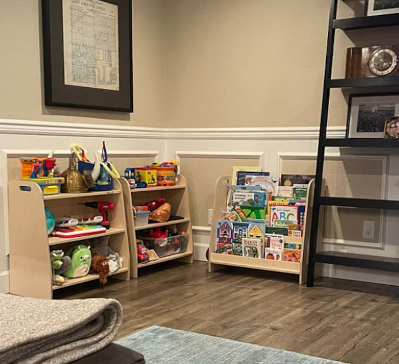 Puede incluir: Dos estanter&iacute;as de madera para juguetes con varios juguetes y libros. Las estanter&iacute;as son de color marr&oacute;n claro y tienen tres estantes cada una. El estante superior de la estanter&iacute;a de la izquierda tiene un coche de juguete rojo, un cami&oacute;n de juguete azul, una pala de juguete amarilla y un casco de juguete plateado. El estante del medio tiene una pelota de juguete azul, un piano de juguete verde y un taladro de juguete plateado. El estante inferior tiene un animal de peluche verde, un animal de peluche marr&oacute;n y un coche de juguete azul. El estante superior de la estanter&iacute;a de la derecha tiene una variedad de libros coloridos. El estante del medio tiene una variedad de libros coloridos. El estante inferior tiene una variedad de libros coloridos. Las estanter&iacute;as est&aacute;n en una habitaci&oacute;n con una pared marr&oacute;n claro y un z&oacute;calo blanco.