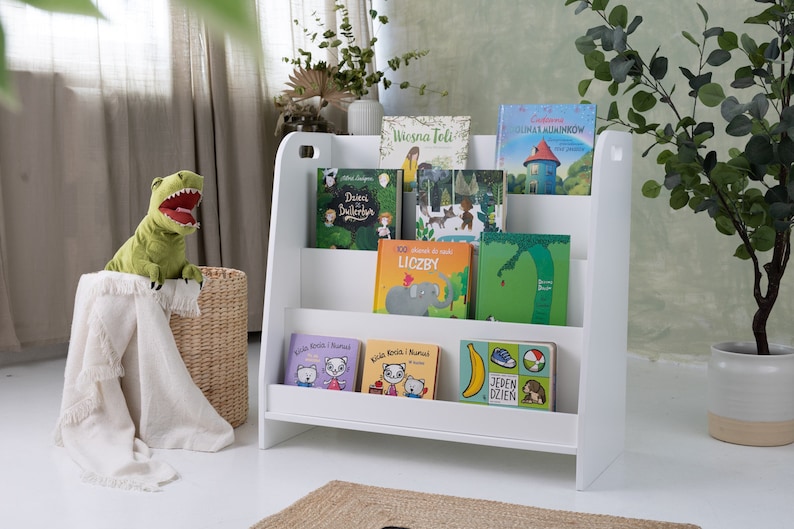 May include: White wooden bookshelf with three shelves, filled with colorful children's books. The books have titles like "Wiosna Feli", "Liczby", and "Jeden Dzien".