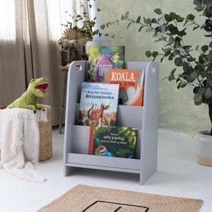 May include: A gray wooden bookshelf with three shelves, holding colorful children's books. A green dinosaur toy sits in a woven basket with a white blanket. A potted plant is to the right.