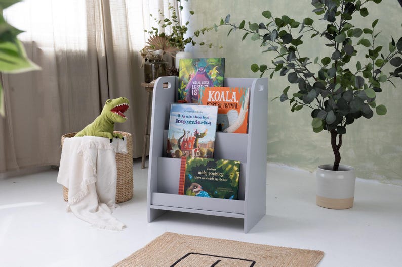 May include: A gray wooden bookshelf with three shelves, holding colorful children's books. A green dinosaur toy sits in a woven basket with a white blanket. A potted plant is to the right.