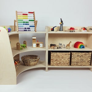 May include: A wooden toy shelf with curved shelves and a straight shelf unit. The shelves are filled with various toys, including a colorful abacus, a toy house, animal figurines, a rainbow arch, and baskets.