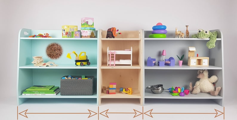 May include: Three wooden shelves, one painted light blue, one natural wood, and one light gray, each with two shelves. The shelves are filled with various toys, including a toy truck, a doll bed, a stacking ring toy, and a stuffed animal.