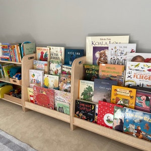 Puede incluir: Una estanter&iacute;a de madera con dos estantes, llena de libros infantiles coloridos. Los libros est&aacute;n organizados por color y tienen t&iacute;tulos en diferentes idiomas, incluyendo ingl&eacute;s, alem&aacute;n y franc&eacute;s. La estanter&iacute;a est&aacute; hecha de madera clara y tiene un dise&ntilde;o sencillo.