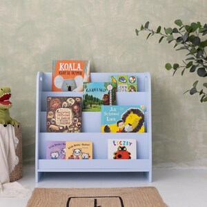 May include: A light blue wooden bookshelf filled with colorful children's books. A green dinosaur toy sits in a woven basket with a white blanket. A potted plant is to the right.