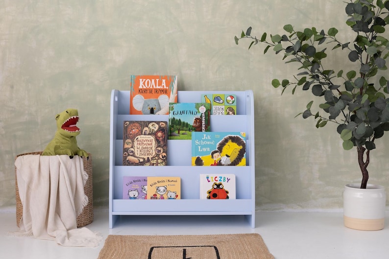 May include: A light blue wooden bookshelf filled with colorful children's books. A green dinosaur toy sits in a woven basket with a white blanket. A potted plant is to the right.