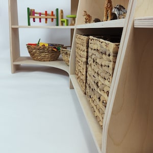 May include: A wooden shelf unit with curved edges and multiple shelves. The shelves are filled with various toys, including a giraffe, a kangaroo, and a set of colorful wooden eggs. There are also two woven baskets on the bottom shelf.