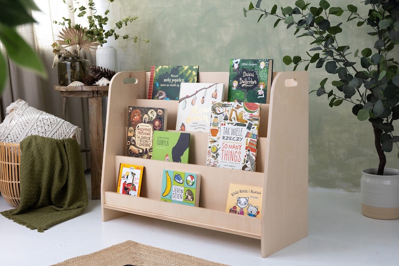 May include: A light wood bookshelf with rounded edges and a handle on the side. The bookshelf has four shelves and is filled with colorful children's books. The books have titles in Polish and English.