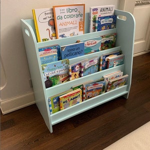 Pu&ograve; includere: Una libreria in legno color azzurro chiaro con quattro ripiani pieni di libri per bambini colorati. I libri hanno titoli in italiano, tra cui "Il mio libro da colorare", "Animali", "Sono un pellicano?", "I Veicoli" e "Colori".