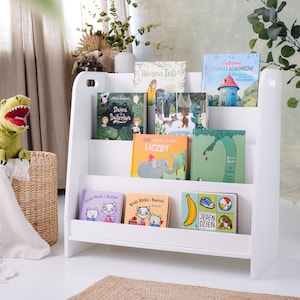 May include: White bookshelf with five shelves, holding a variety of children's books. The books have colorful covers with illustrations and titles in Polish. A green dinosaur plush toy is sitting in a wicker basket.