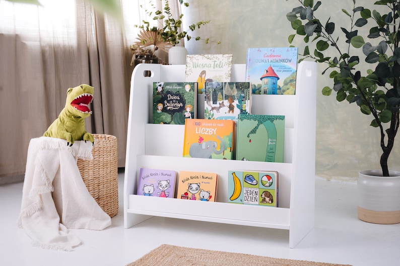 May include: White bookshelf with five shelves, holding a variety of children's books. The books have colorful covers with illustrations and titles in Polish. A green dinosaur plush toy is sitting in a wicker basket.