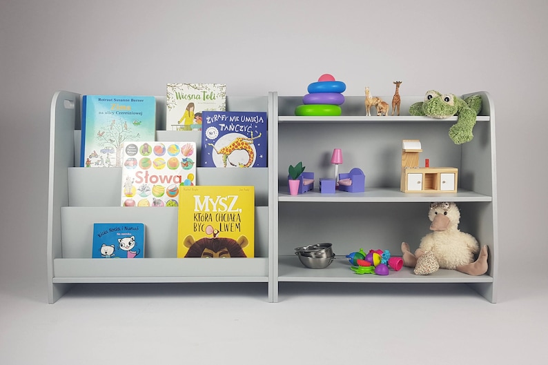 Pu&ograve; includere: Una libreria in legno grigia con due ripiani su ogni lato. Il lato sinistro ha un organizzatore di libri con diversi libri per bambini colorati. Il lato destro ha una variet&agrave; di giocattoli, tra cui una rana di peluche, un'anatra di peluche, una cucina giocattolo in legno e una pila di anelli colorati.