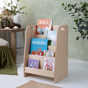 May include: A light-colored wooden bookshelf with multiple shelves, holding several children's books with colorful covers. The bookshelf has a simple design with cut-out handles. The books feature illustrations of animals and other characters.