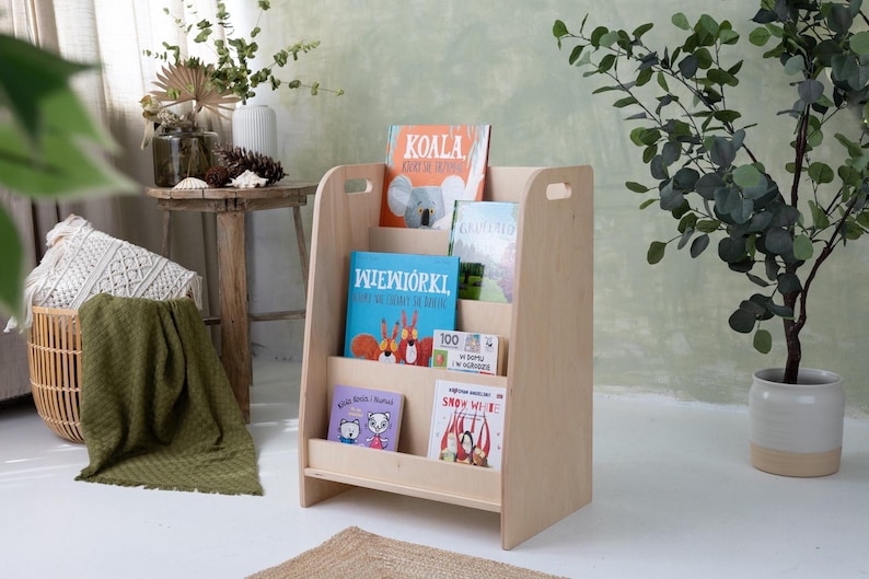 May include: A light-colored wooden bookshelf with multiple shelves, holding several children's books with colorful covers. The bookshelf has a simple design with cut-out handles. The books feature illustrations of animals and other characters.