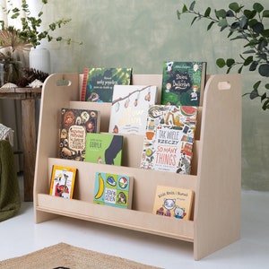May include: A light wood children's bookshelf with rounded edges and handles on the sides. The bookshelf is filled with colorful children's books with various titles in Polish and English. The bookshelf is standing on a white floor.