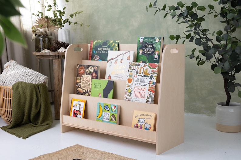 May include: A light wood children's bookshelf with rounded edges and handles on the sides. The bookshelf is filled with colorful children's books with various titles in Polish and English. The bookshelf is standing on a white floor.