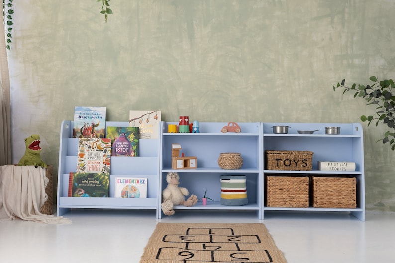 Puede incluir: Una estanter&iacute;a de color azul claro con tres estantes, que presenta una variedad de libros para ni&ntilde;os, juguetes y cestas de almacenamiento. La estanter&iacute;a est&aacute; contra una pared texturizada de color verde claro.