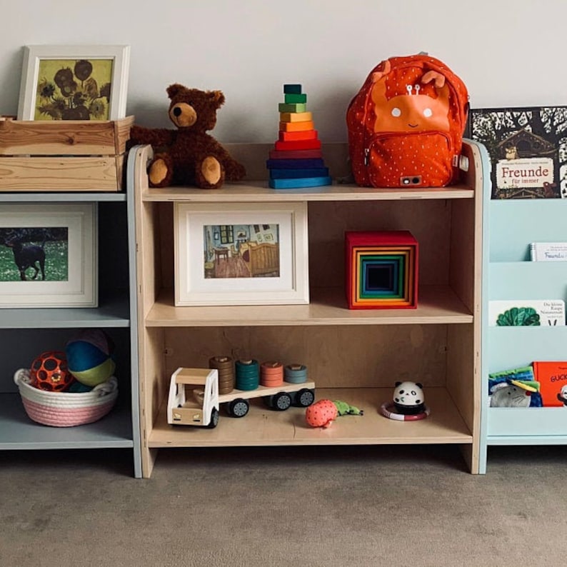 Puede incluir: Una estanter&iacute;a de madera con tres estantes, que presenta una variedad de juguetes y libros para ni&ntilde;os. El estante superior tiene una mochila roja con una cara blanca, un juguete de apilar arco&iacute;ris y un oso de peluche marr&oacute;n. El estante del medio tiene una imagen enmarcada, un juguete de apilar arco&iacute;ris y un cami&oacute;n de juguete. El estante inferior tiene una cesta de pelotas, un juguete de panda y un libro titulado "Freunde f&uuml;r immer".