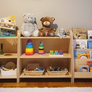 May include: Three wooden shelves with various toys and books for children. The top shelf has two stuffed animals, a gray bear and a brown bear. The middle shelf has a rainbow stacking toy, a wooden stacking toy, and a set of colorful nesting cups. The bottom shelf has a green plastic bin filled with toys, a white basket with wooden toys, and two wicker baskets with natural materials. The right shelf has a collection of children's books with colorful covers.