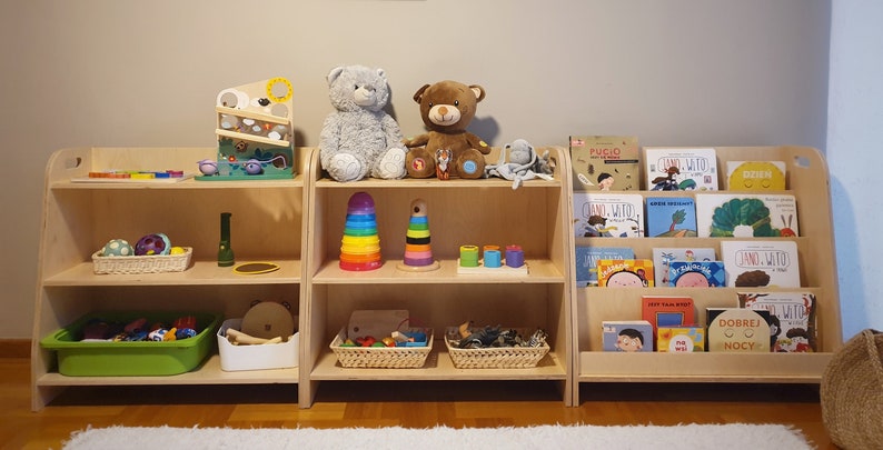 May include: Three wooden shelves with various toys and books for children. The top shelf has two stuffed animals, a gray bear and a brown bear. The middle shelf has a rainbow stacking toy, a wooden stacking toy, and a set of colorful nesting cups. The bottom shelf has a green plastic bin filled with toys, a white basket with wooden toys, and two wicker baskets with natural materials. The right shelf has a collection of children's books with colorful covers.