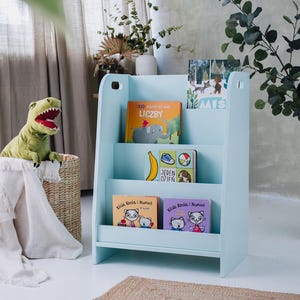 May include: A light blue wooden bookshelf with three shelves, filled with colorful children's books. A green dinosaur toy sits in a woven basket next to the bookshelf. A white blanket is draped over the basket.
