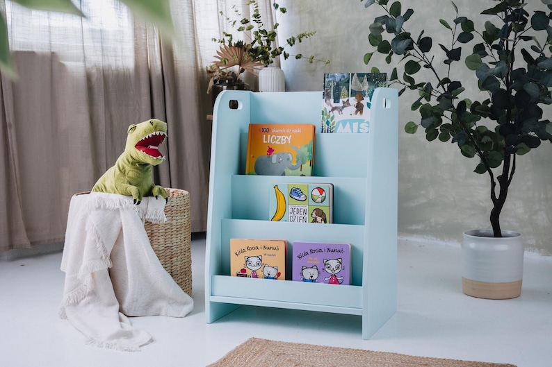 May include: A light blue wooden bookshelf with three shelves, filled with colorful children's books. A green dinosaur toy sits in a woven basket next to the bookshelf. A white blanket is draped over the basket.