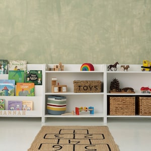 Puede incluir: Estanter&iacute;a blanca con tres estantes, con un arco&iacute;ris, una caja de juguetes etiquetada como "TOYS" y una variedad de juguetes, incluyendo un cami&oacute;n de juguete, un coche de juguete y un dinosaurio de juguete. La estanter&iacute;a est&aacute; etiquetada como "HANNAH" en el estante inferior.
