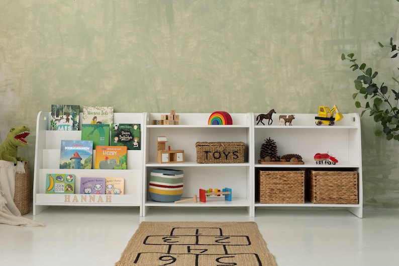 Puede incluir: Estanter&iacute;a blanca con tres estantes, con un arco&iacute;ris, una caja de juguetes etiquetada como "TOYS" y una variedad de juguetes, incluyendo un cami&oacute;n de juguete, un coche de juguete y un dinosaurio de juguete. La estanter&iacute;a est&aacute; etiquetada como "HANNAH" en el estante inferior.
