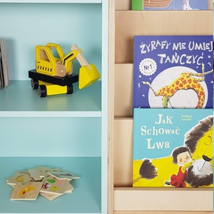 May include: A light blue bookshelf with three shelves, a wooden book holder with three slots, and various children's books. The bookshelf is filled with toys, including a yellow toy excavator, a set of wooden blocks, and a puzzle.