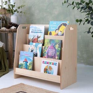 May include: A light-colored wooden bookshelf with multiple shelves, holding several children's books with colorful covers. The bookshelf has a simple, angled design and is placed in a bright room with plants and a rug.