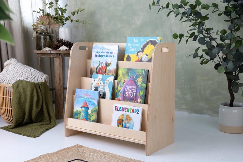 May include: A light-colored wooden bookshelf with multiple shelves, holding several children's books with colorful covers. The bookshelf has a simple, angled design and is placed in a bright room with plants and a rug.