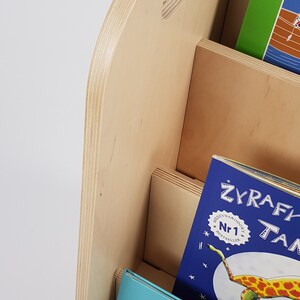 May include: A wooden bookshelf with three shelves, each holding a different children's book. The top shelf has a green book with a picture of a boat on the cover. The middle shelf has a blue book with a picture of a giraffe on the cover. The bottom shelf has a blue book with a picture of a lion on the cover. The bookshelf is made of light wood and has a handle on the top.