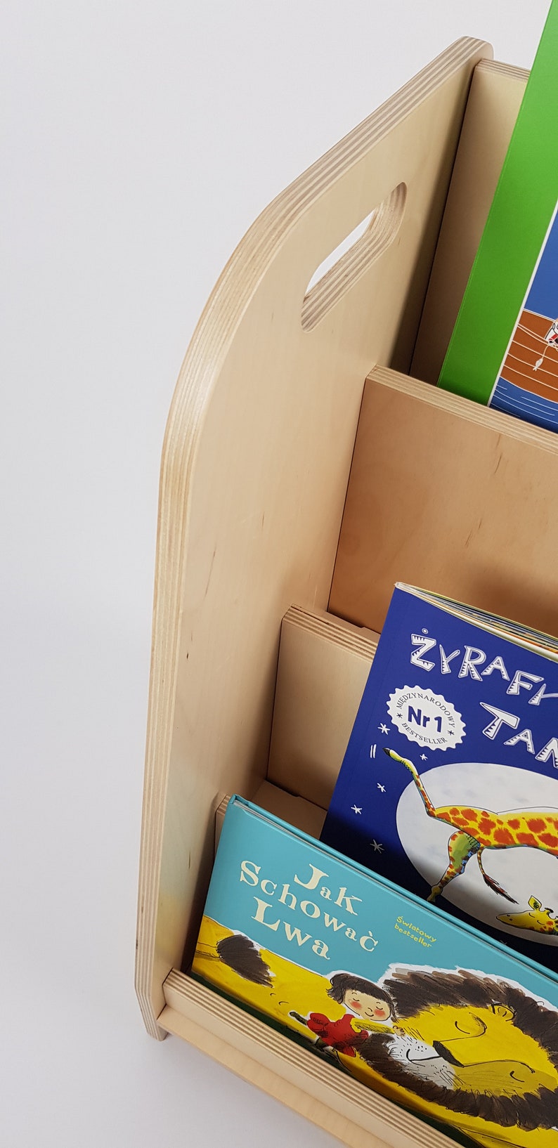 May include: A wooden bookshelf with three shelves, each holding a different children's book. The top shelf has a green book with a picture of a boat on the cover. The middle shelf has a blue book with a picture of a giraffe on the cover. The bottom shelf has a blue book with a picture of a lion on the cover. The bookshelf is made of light wood and has a handle on the top.