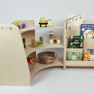 May include: A wooden bookshelf with three shelves, a curved front, and a handle on the side. The shelves are filled with children's books, toys, and a basket. The bookshelf is made of natural wood and has a light brown finish.