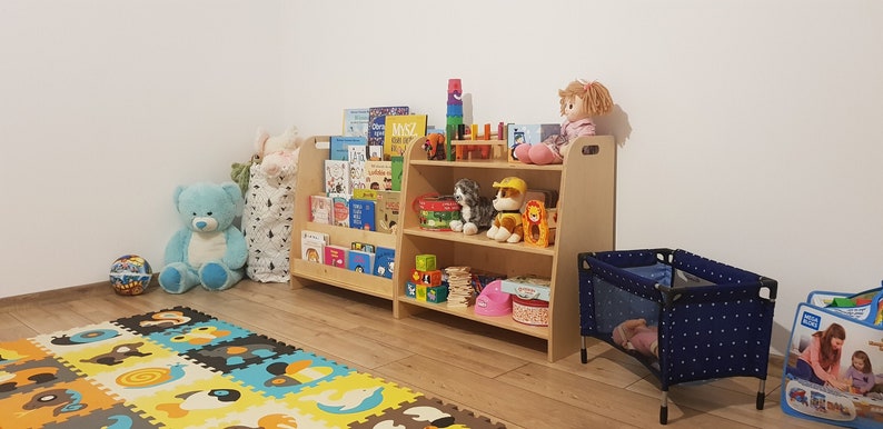 May include: A wooden bookshelf with two shelves filled with books, toys, and stuffed animals. The bookshelf is in a playroom with a blue and white playpen and a colorful foam play mat on the floor.